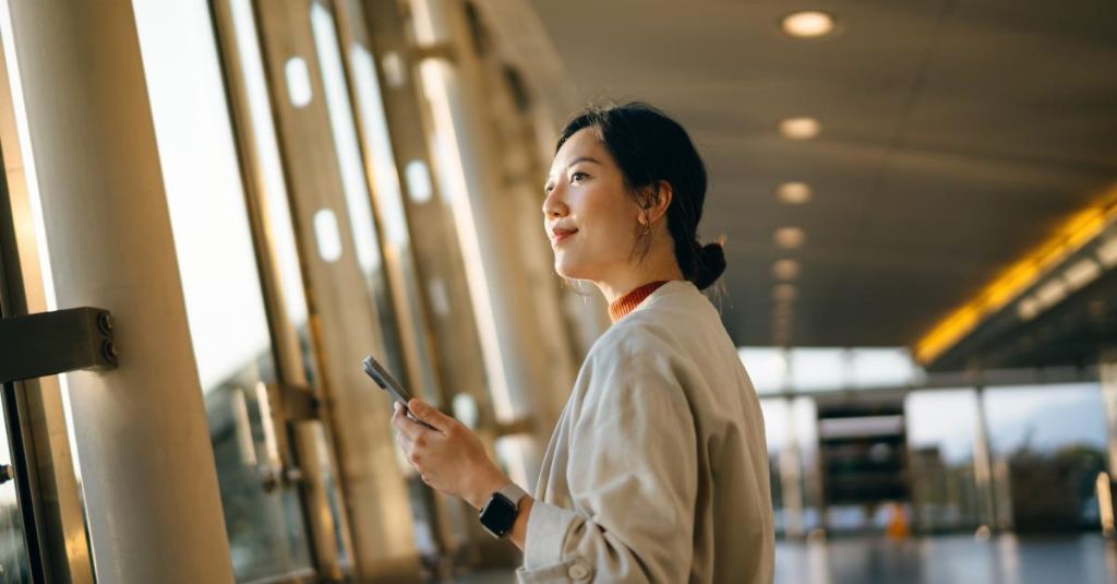 Young woman looking through a big window while holds her smartphone